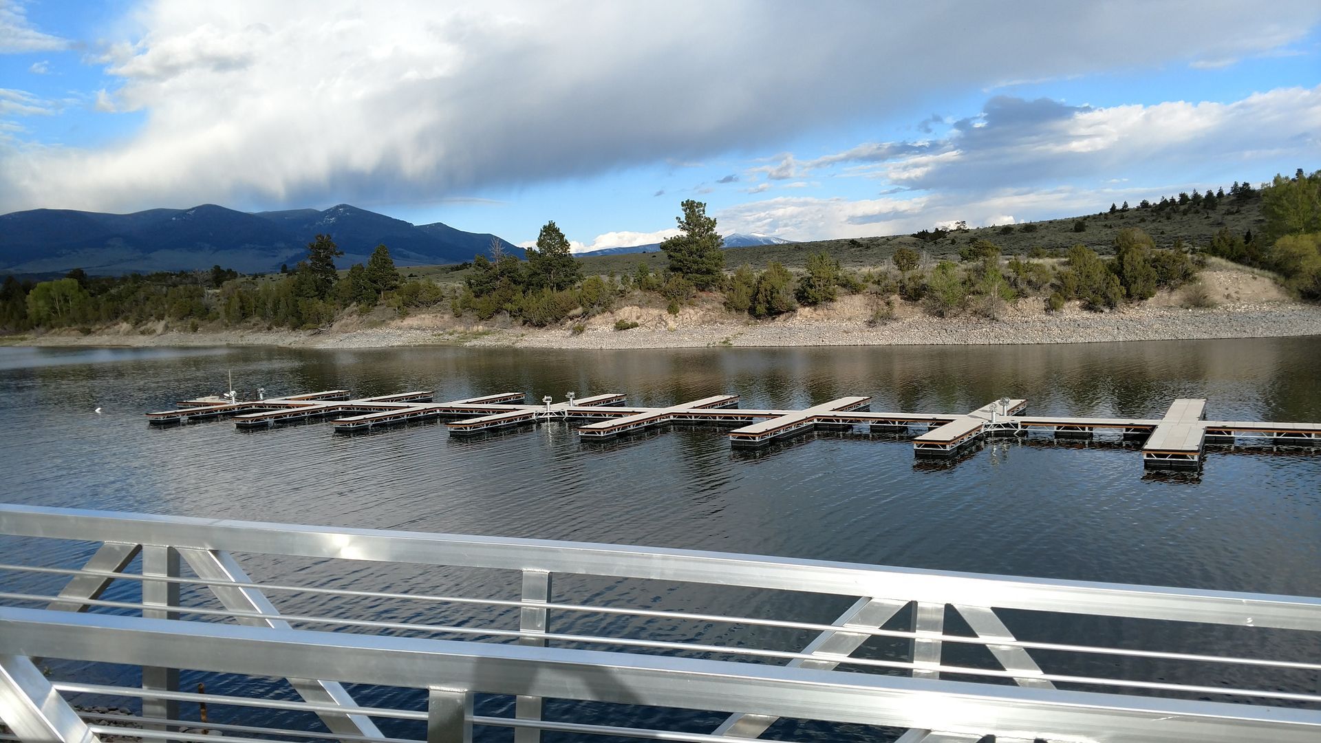 Docked boats on a calm lake with mountains and cloudy sky, viewed from a bridge.