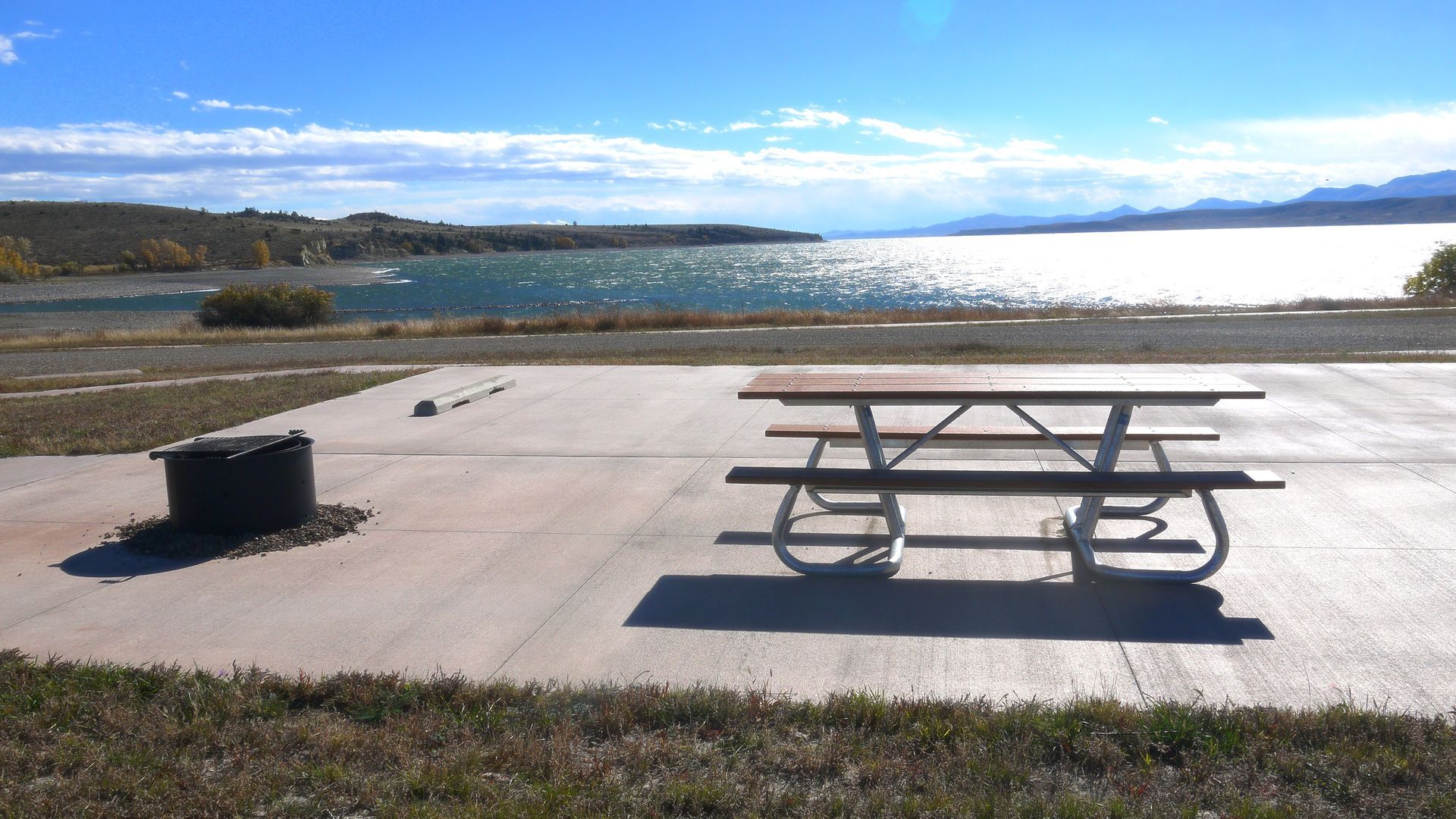 Picnic table on a sunny waterfront patio with a lake and distant hills in the background