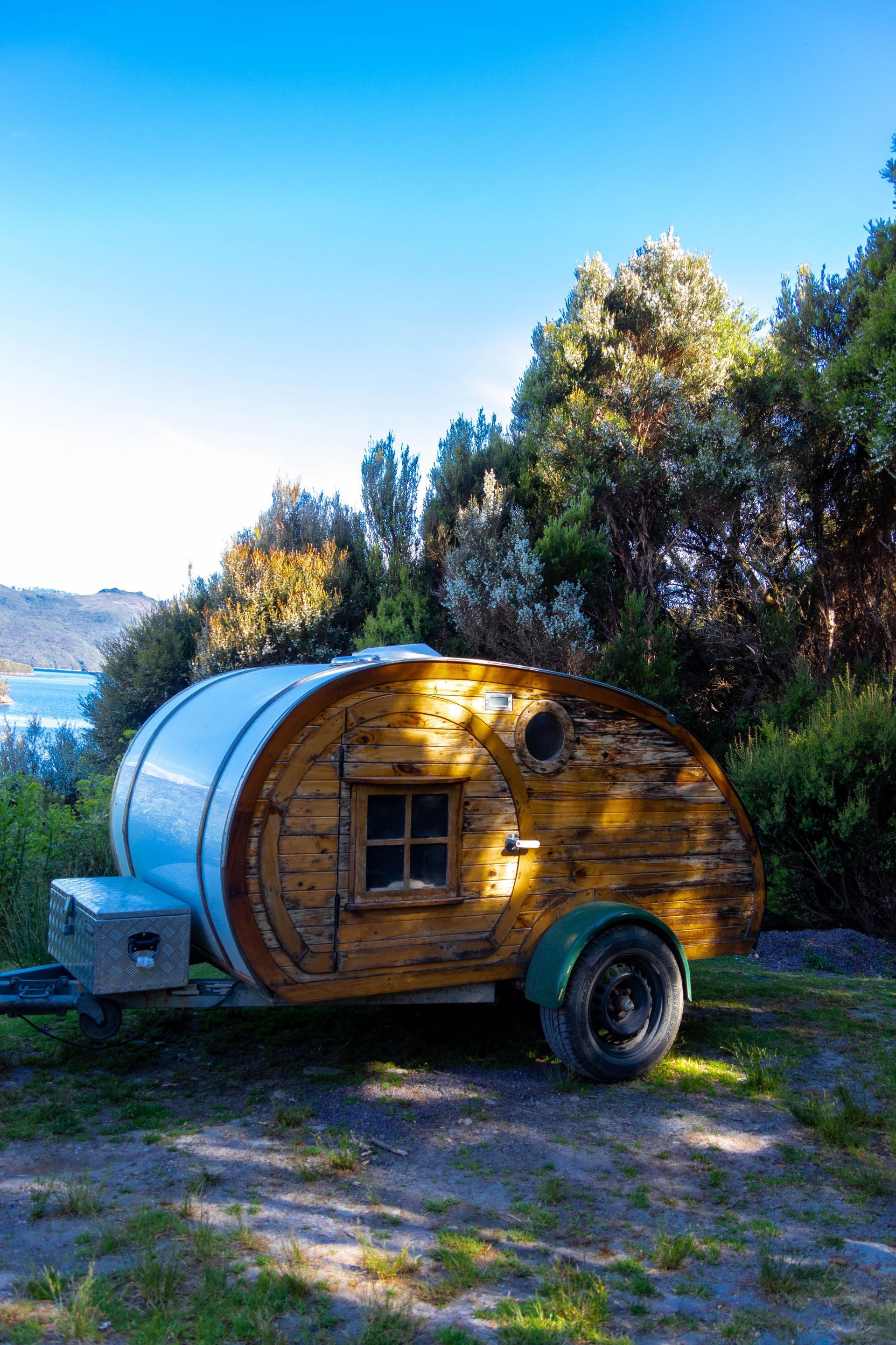 Wooden teardrop camper trailer parked on grass under trees in a sunny rural landscape