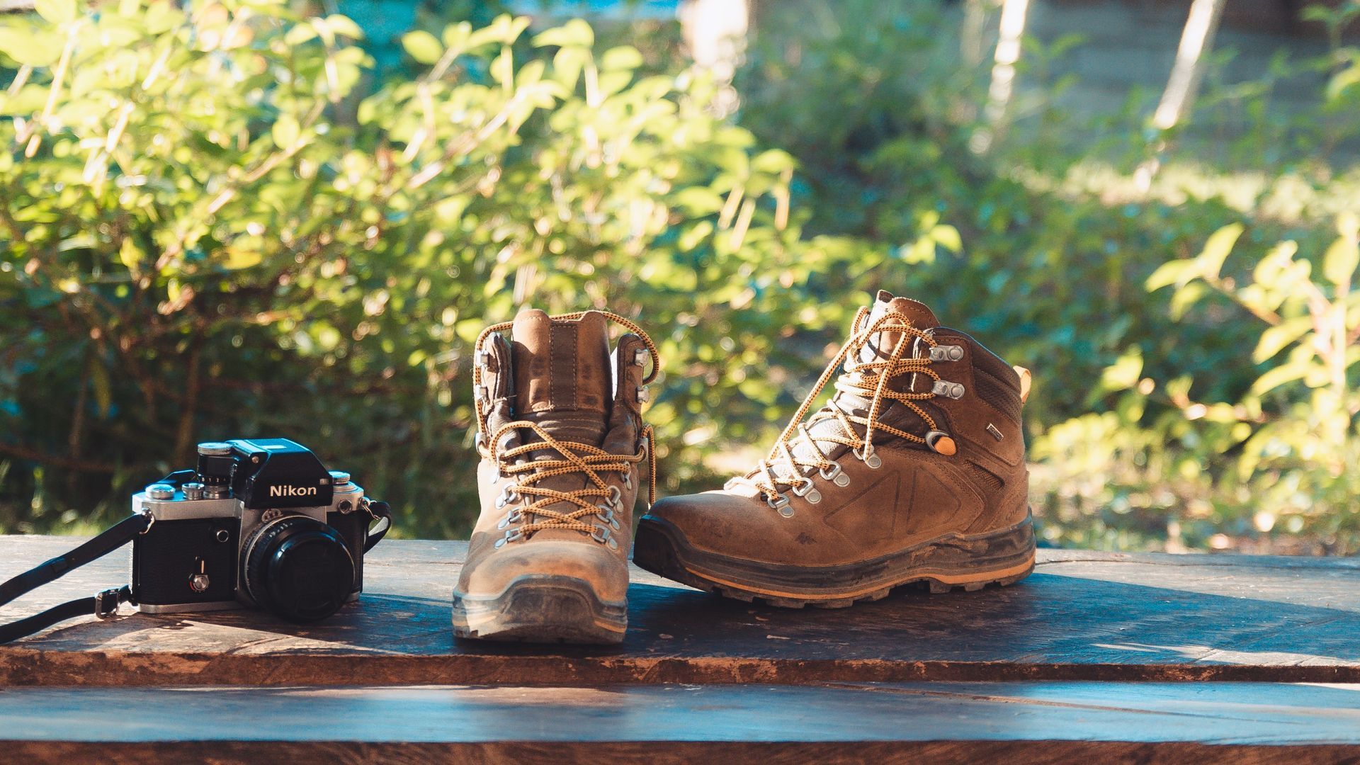 Brown hiking boots and a camera on a wooden bench outdoors in sunlight