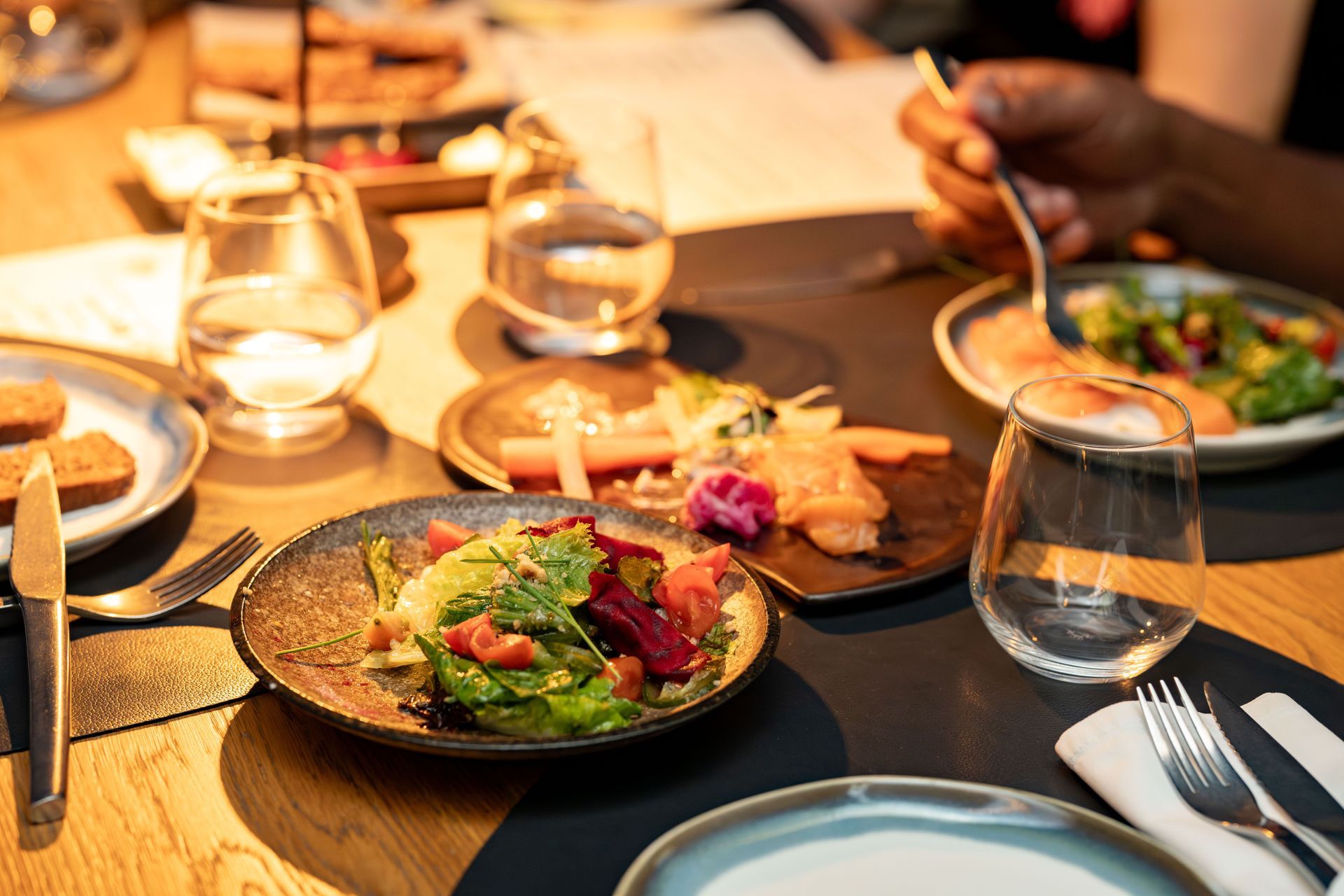 Warm-lit dining table with plated salads, glasses, candles, and hands serving food