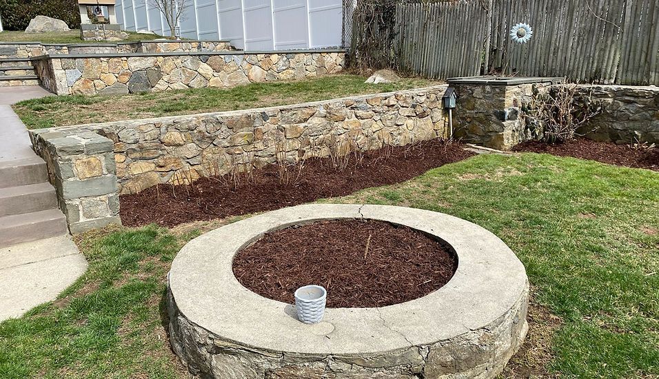 A round stone-walled garden bed with dark mulch and a small planter sits in a backyard near tiered stone retaining walls.
