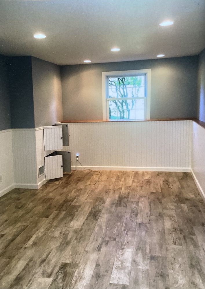 A room with gray walls, white wainscoting, a centered window, and wood-look flooring with a small white cabinet.