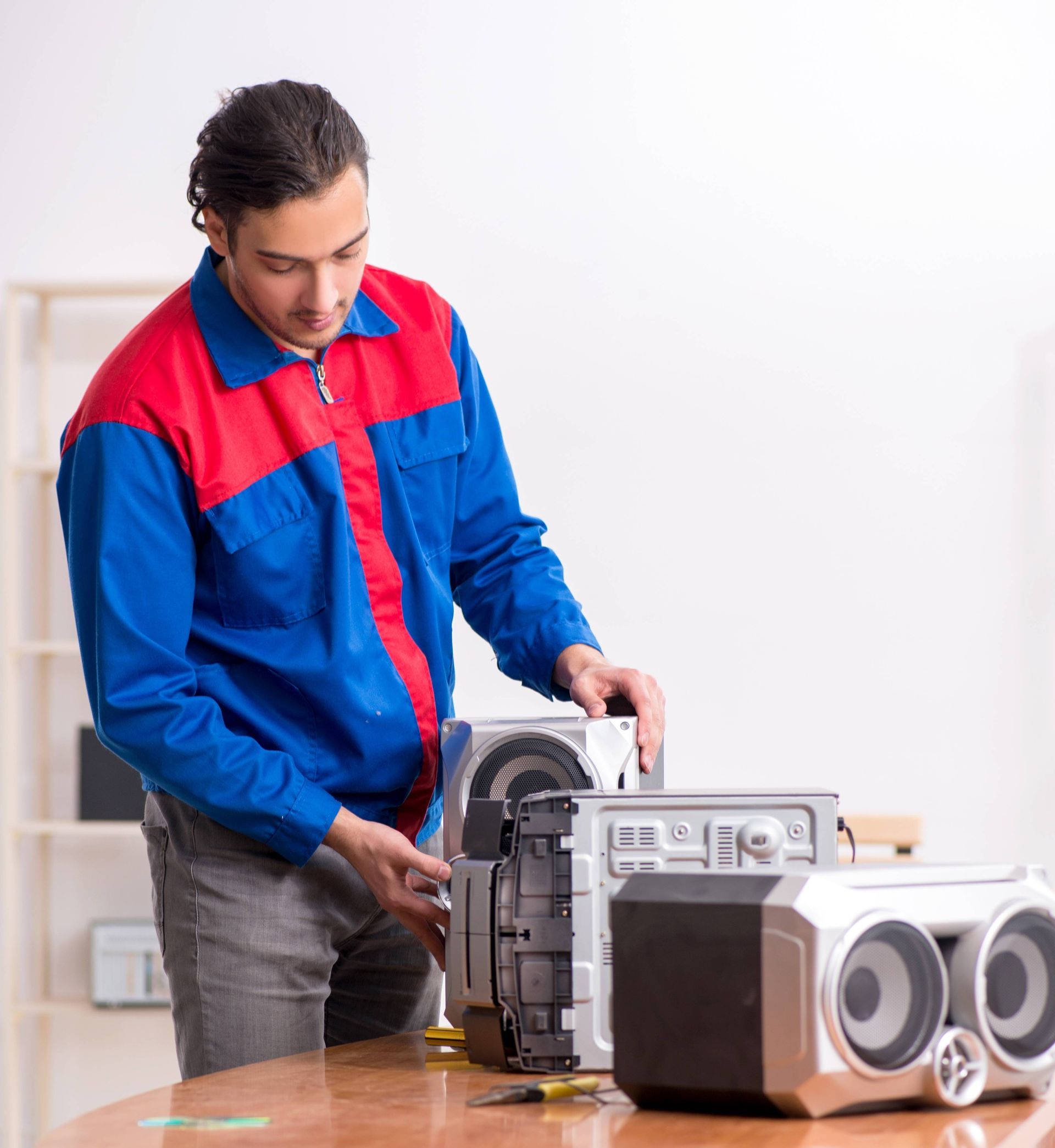 A technician wearing a blue and red jacket repairs an audio system on a wooden table.