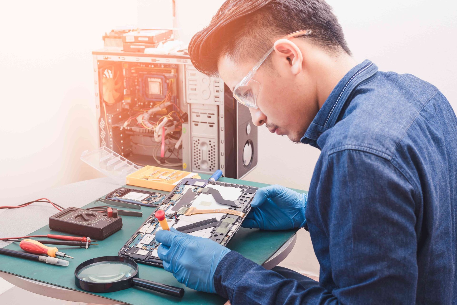 A technician in a denim shirt and blue gloves repairs an electronic device on a workbench with tools and a computer case.