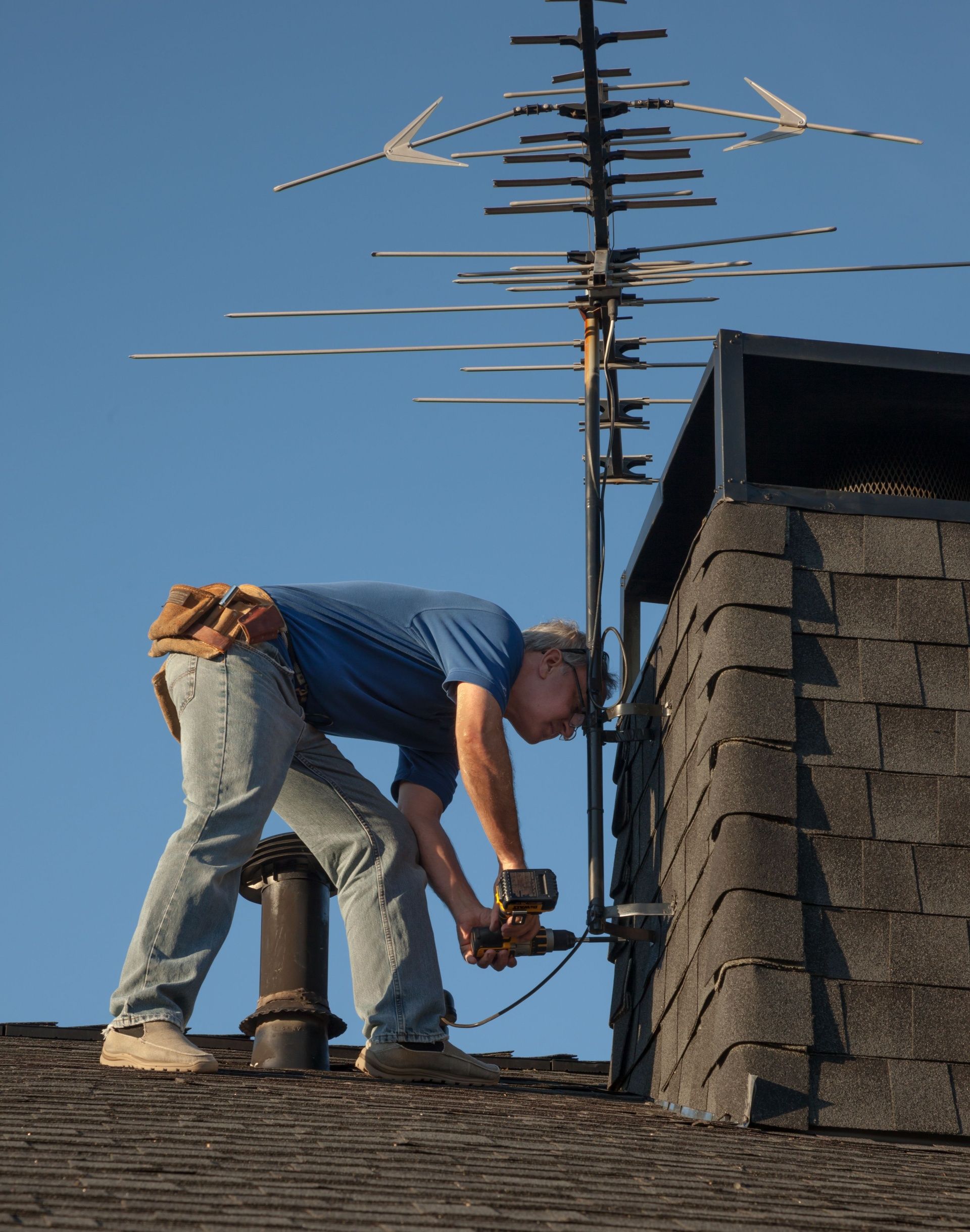 A person working on a roof, using a power drill to attach a tall television antenna to a chimney.