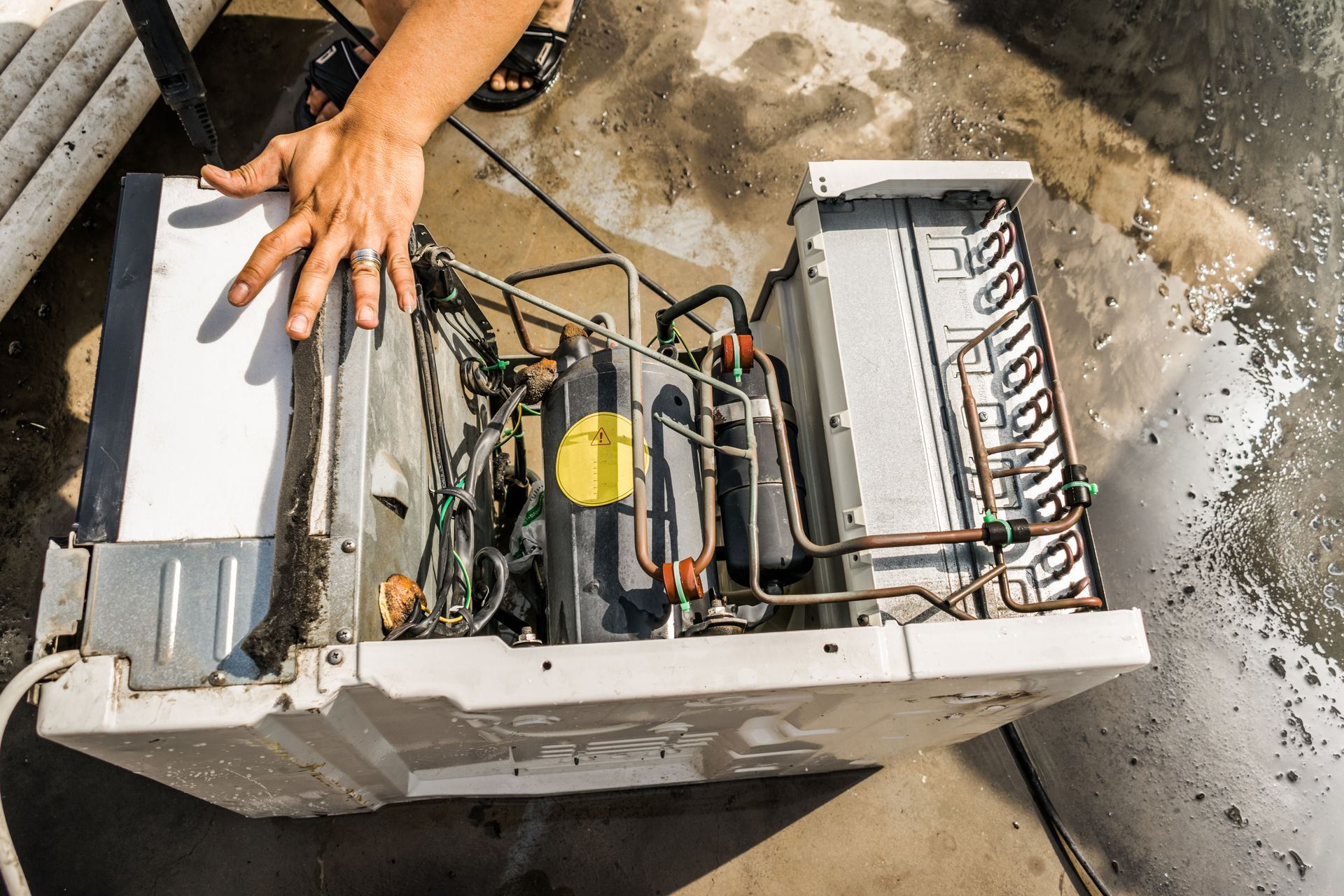 A person’s hand rests on the casing of an open window air conditioning unit undergoing maintenance on a concrete surface.