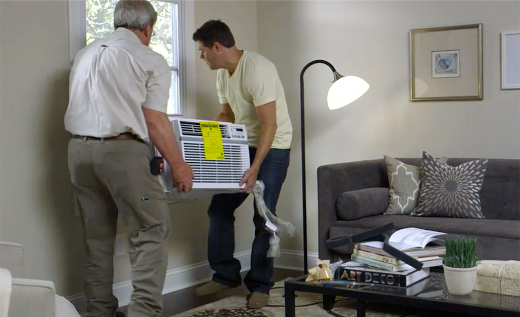 Two people lift a window air conditioner unit toward an open window in a living room.