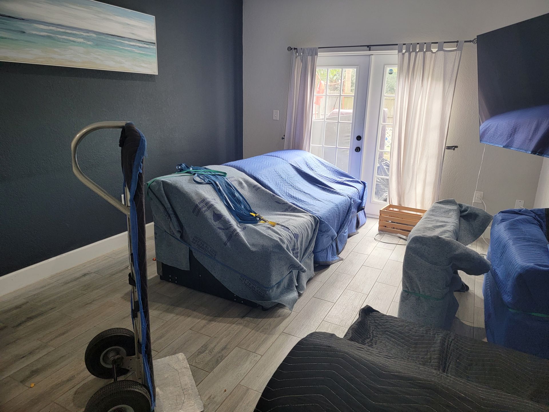 Messy bedroom with blue-covered beds, sheer curtains, and sunlight by the balcony doors