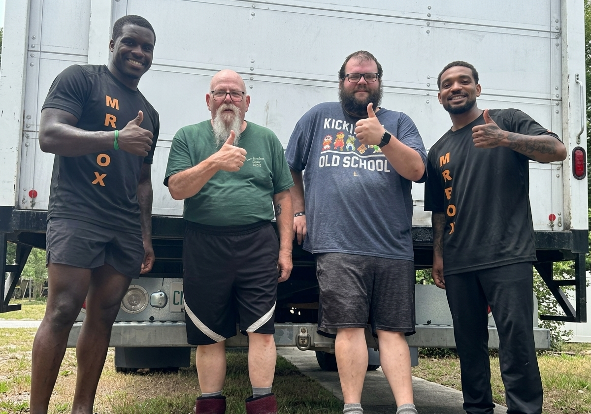 Four people standing and smiling in front of a truck, posing outdoors on wet pavement.