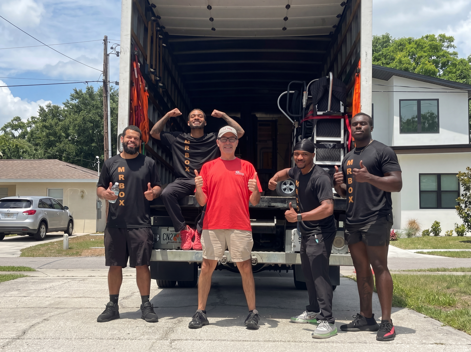 Five people posing at the back of a large moving truck in a neighborhood street.
