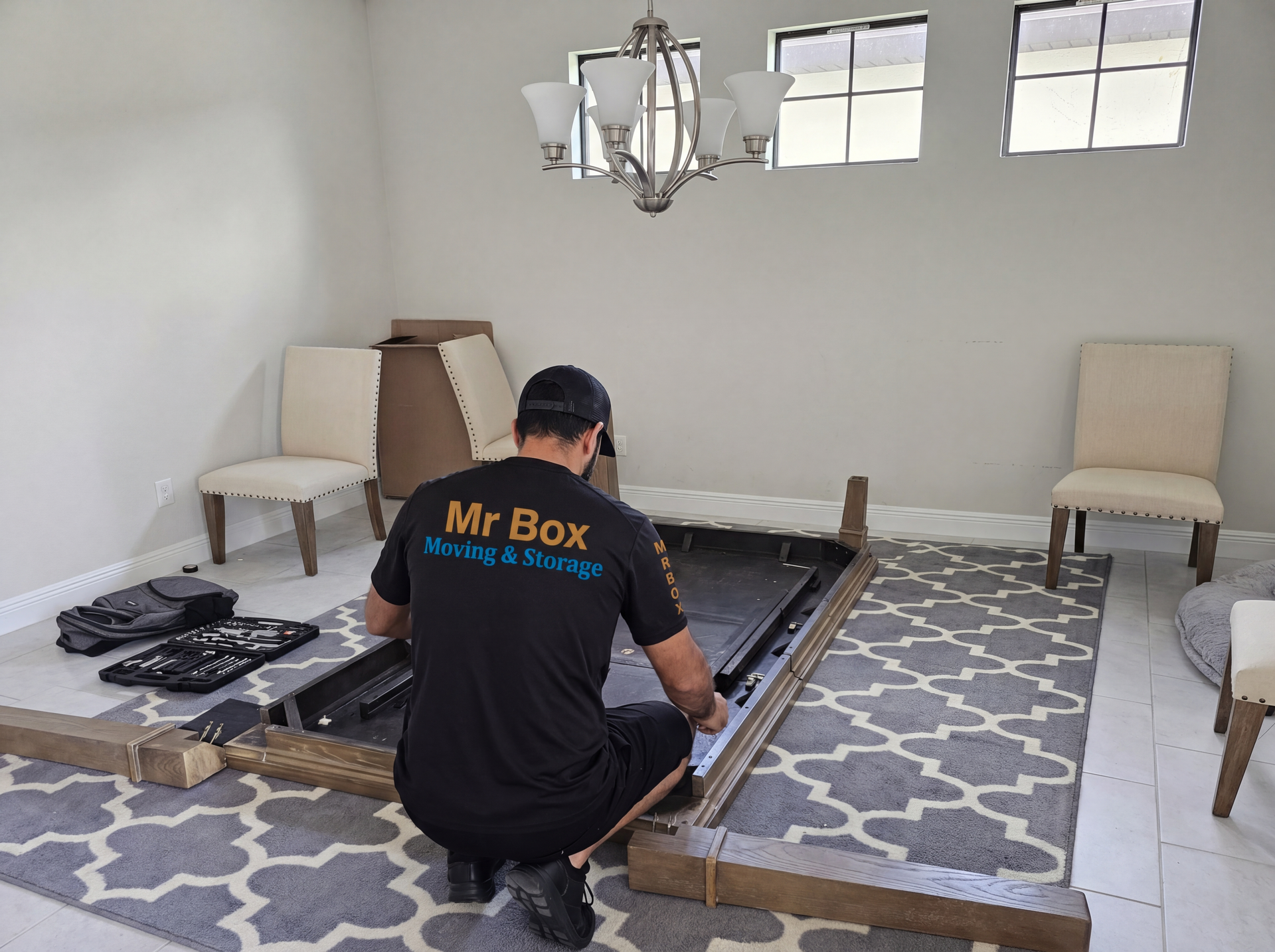 Mover assembling furniture in a bright room with boxes, chairs, and a rug on the floor