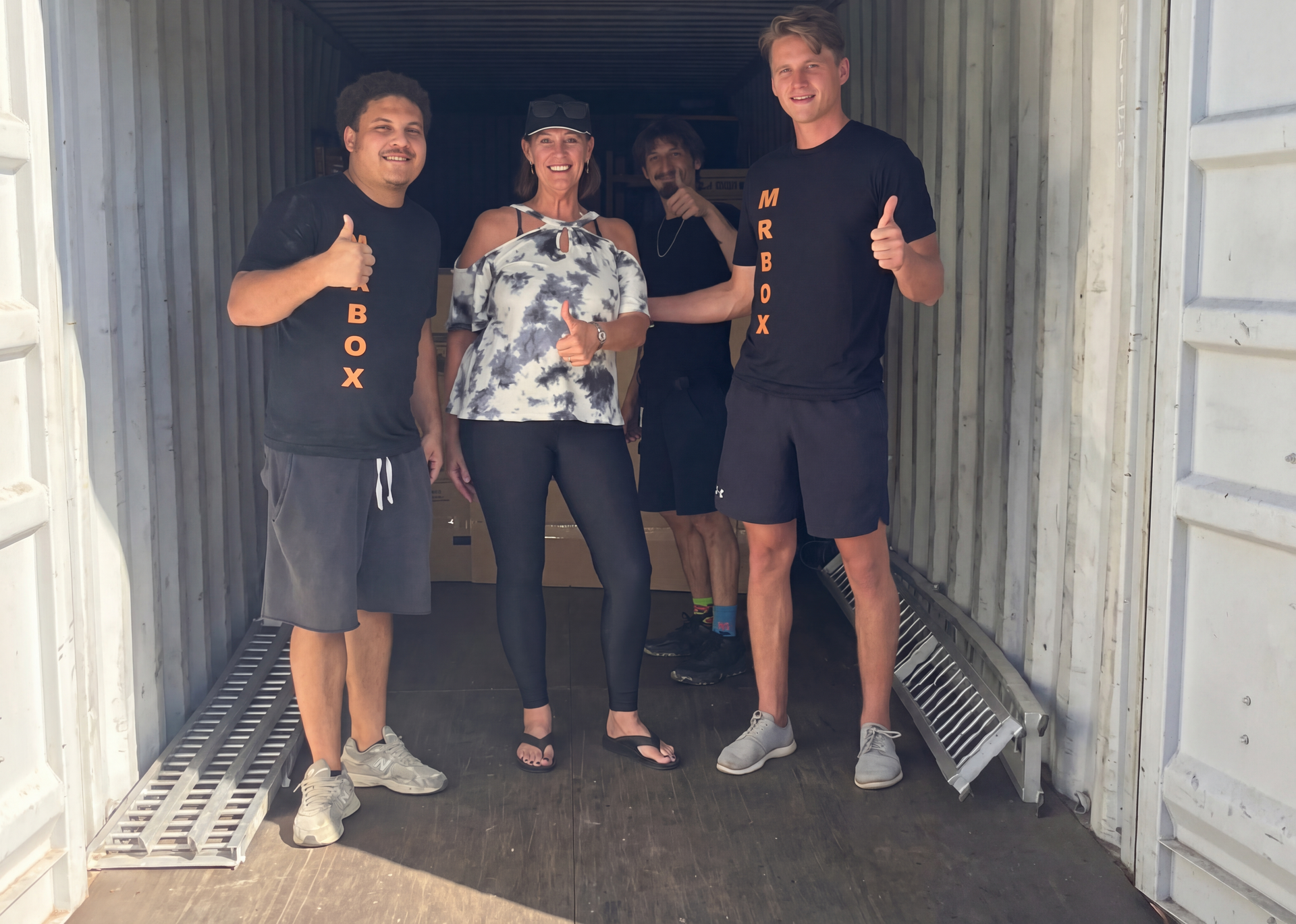 Three people standing in a shipping container doorway, smiling and giving thumbs-up.