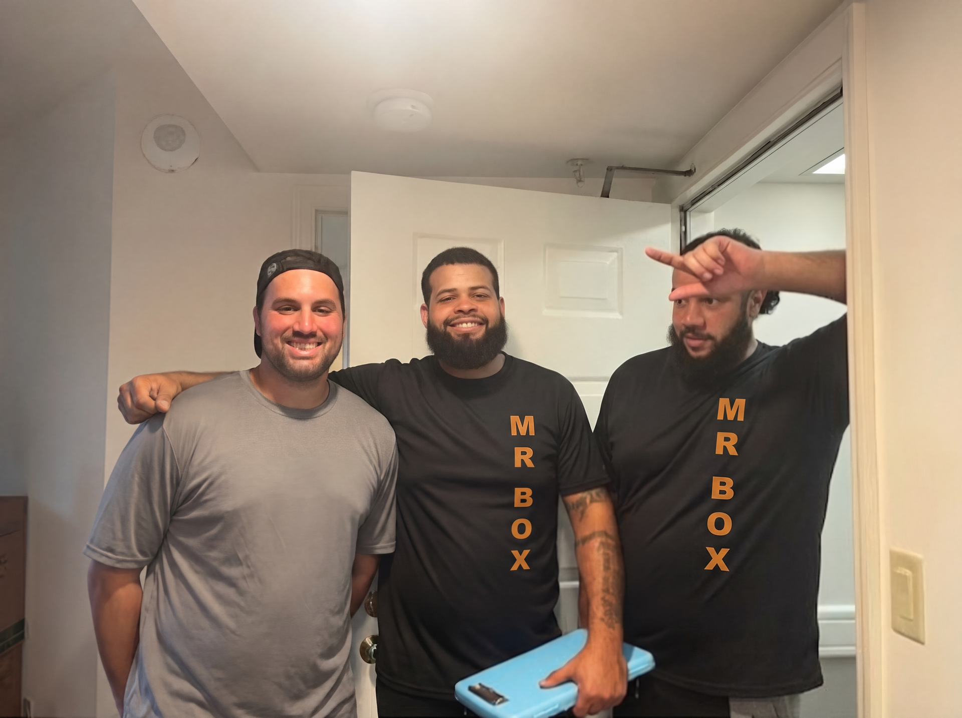 Three smiling men in black “MR BOX” shirts stand indoors, with one flashing a peace sign.