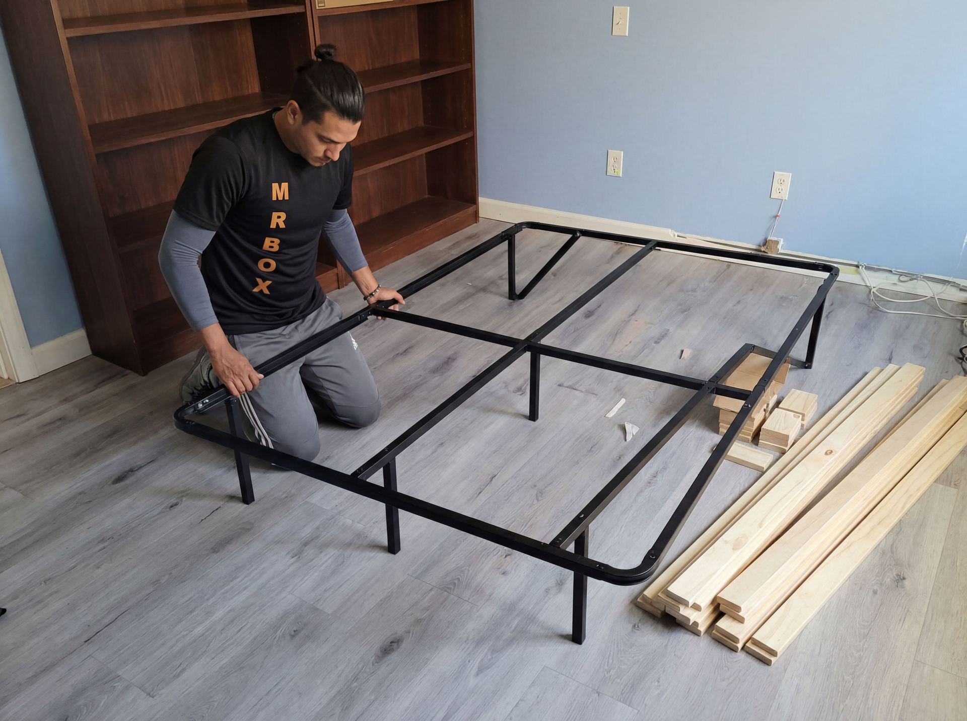 Person assembling a black metal bed frame on a floor beside stacked wooden planks and shelving