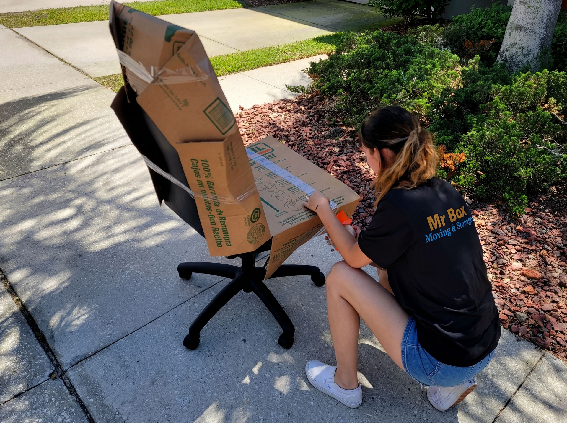Woman kneeling beside a cardboard chair outdoors, working on it with a tool.