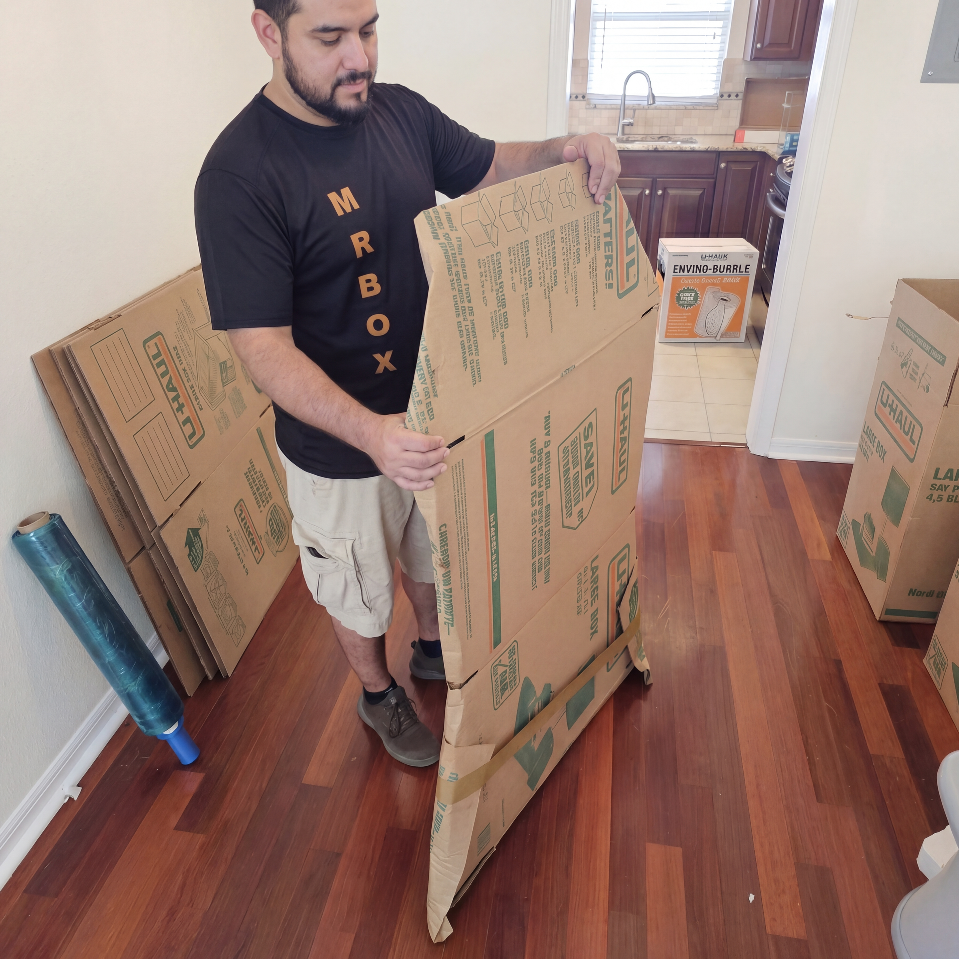 Man assembling a large cardboard moving box in a hardwood-floored room.