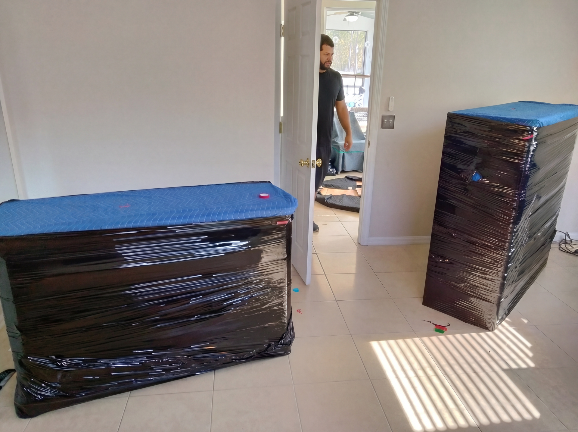 Wrapped furniture in a room; a person stands in the doorway with sunlight on the floor.