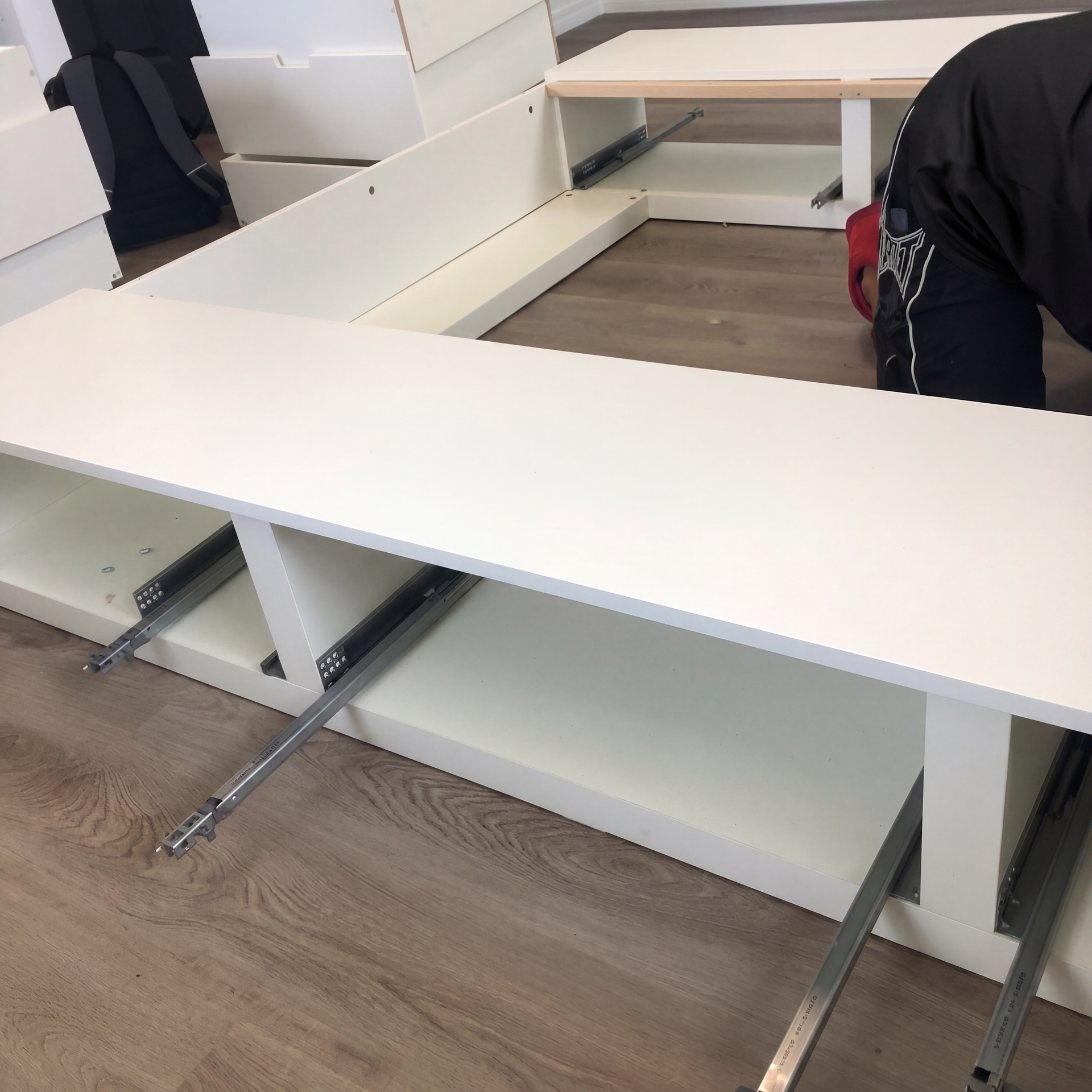 White office desk with drawers and metal supports, viewed from above on a wood floor; person partly visible at right.