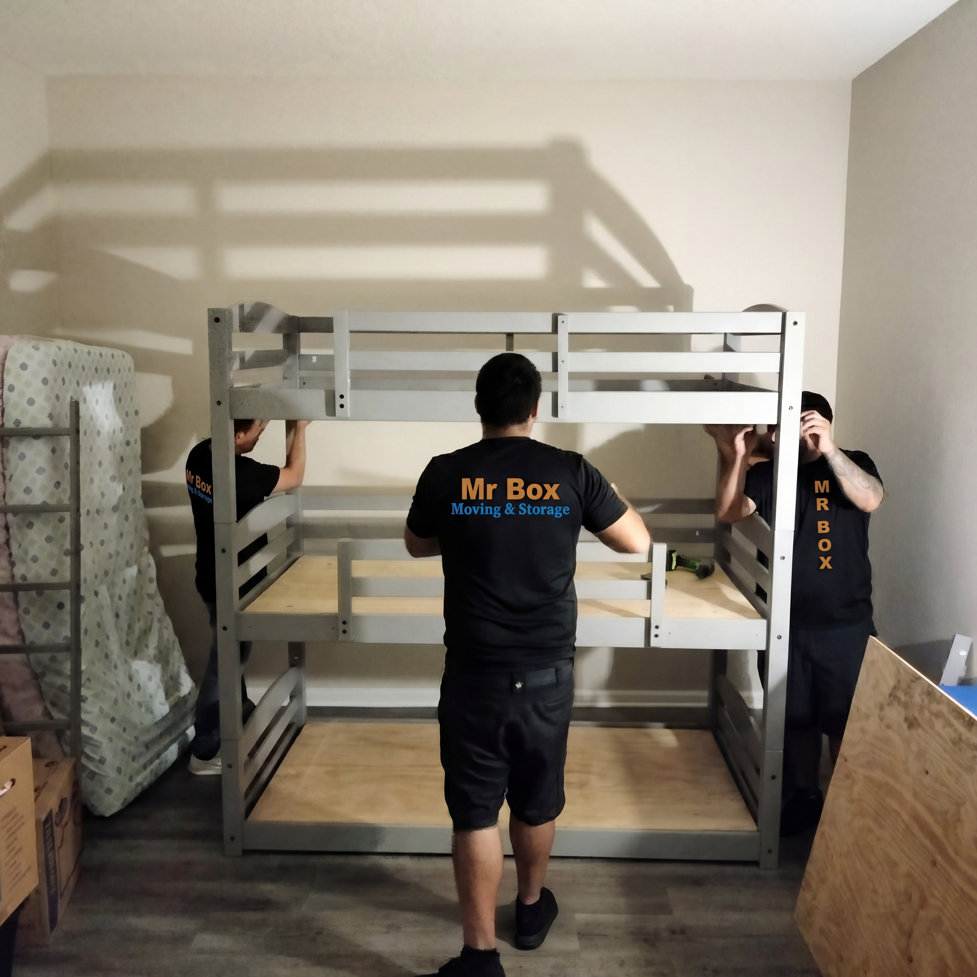 Movers assembling a metal bunk bed frame in a bare room, with mattress and plywood nearby.