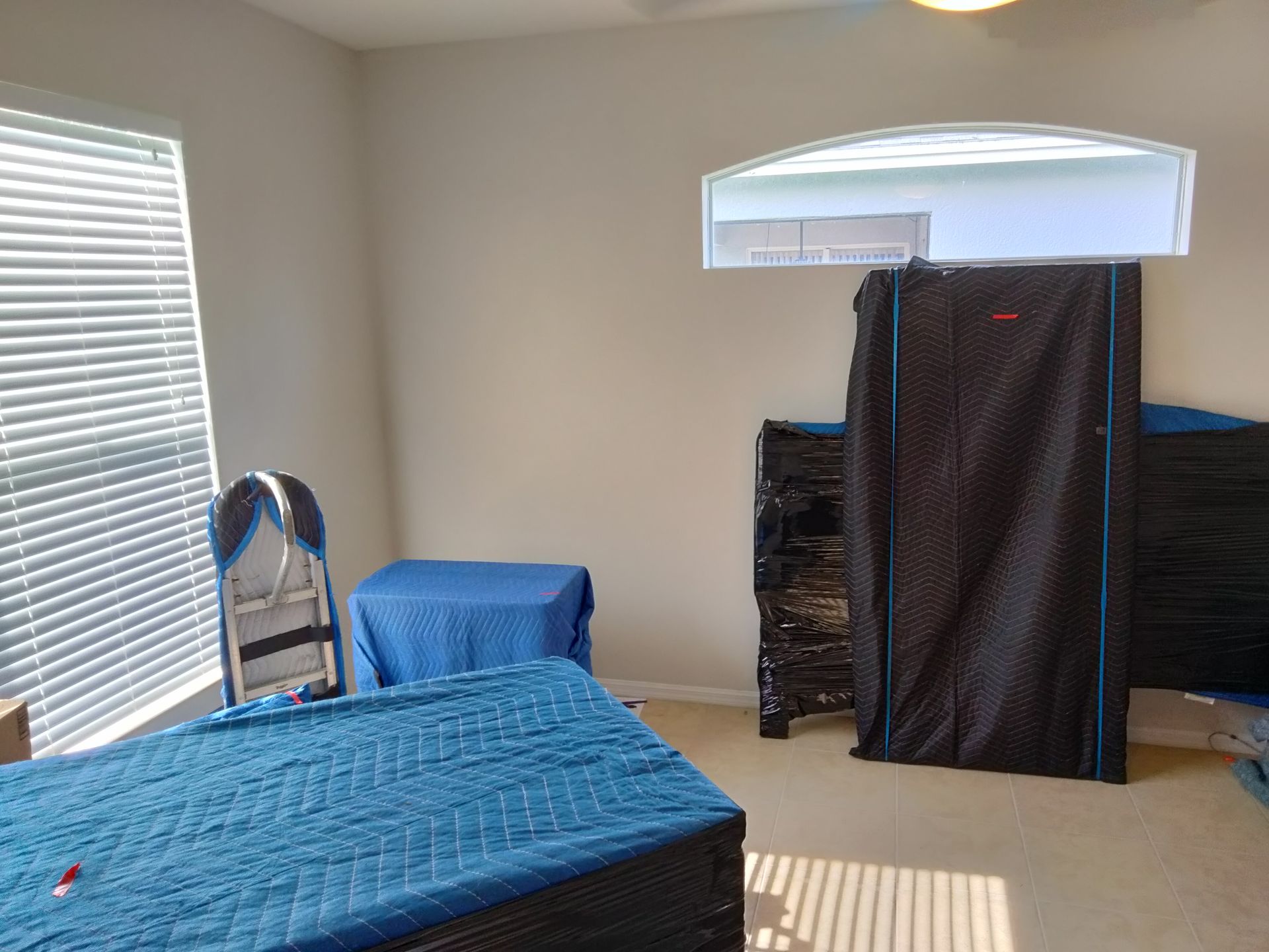 Bedroom with a blue bedspread, window blinds, and a black privacy tent near the corner.