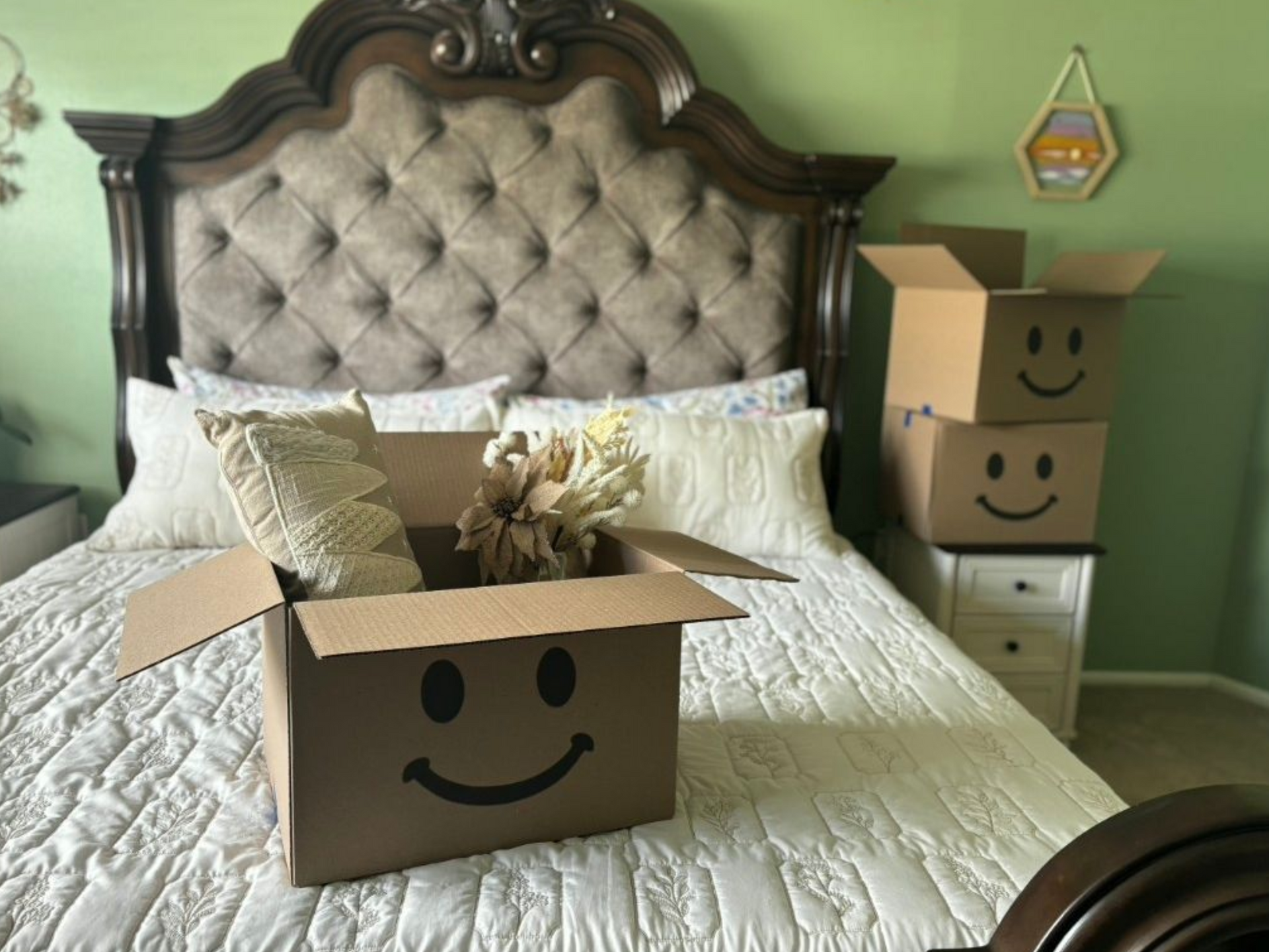 Two smiling cardboard boxes in a green bedroom, one on a bed and one on a nightstand.