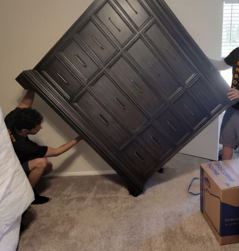 Two people pushing a large dark dresser tilted dangerously in a bedroom during a move