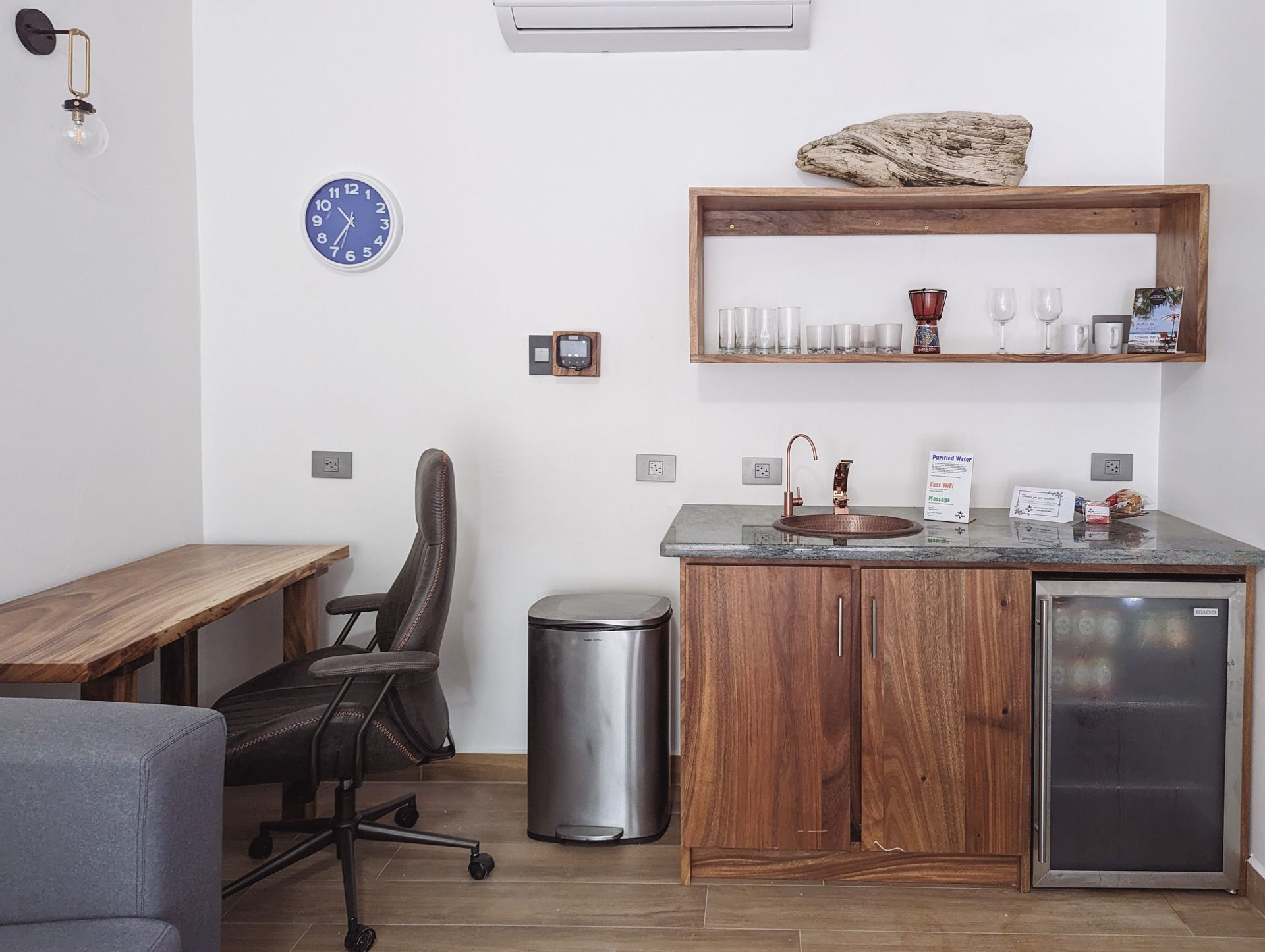 A room with a desk , chair , trash can and a clock on the wall.