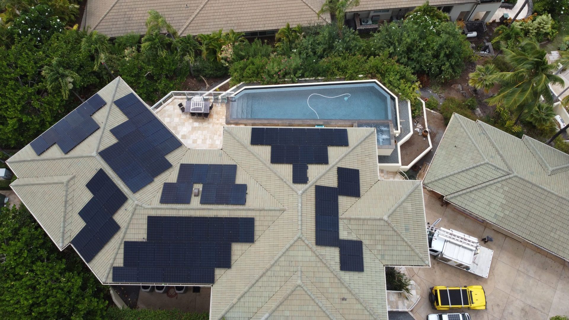 Aerial view of a house roof covered with dark solar panels beside a backyard pool.