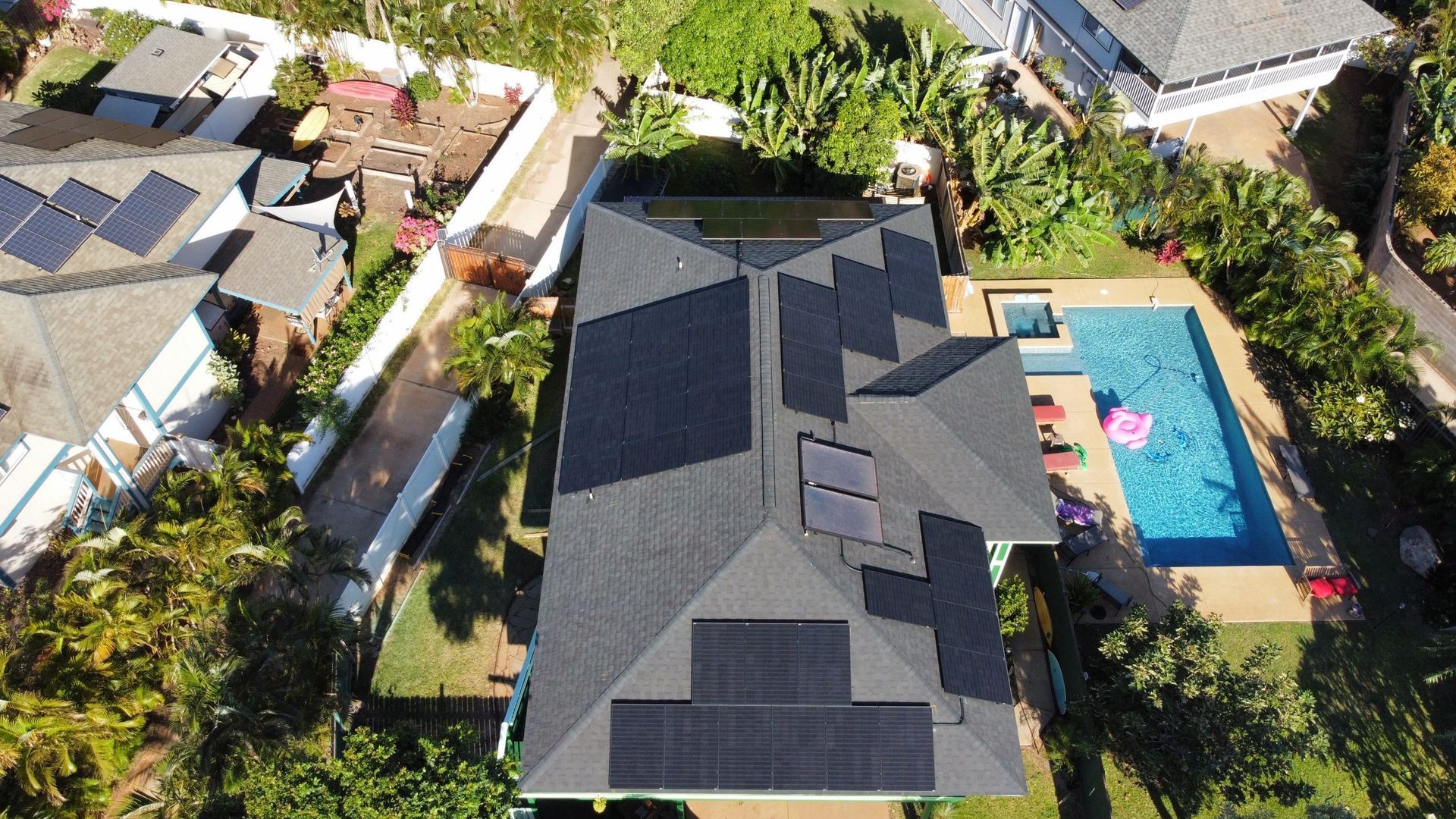 Aerial view of a house with a dark roof beside a backyard swimming pool and neighboring homes.