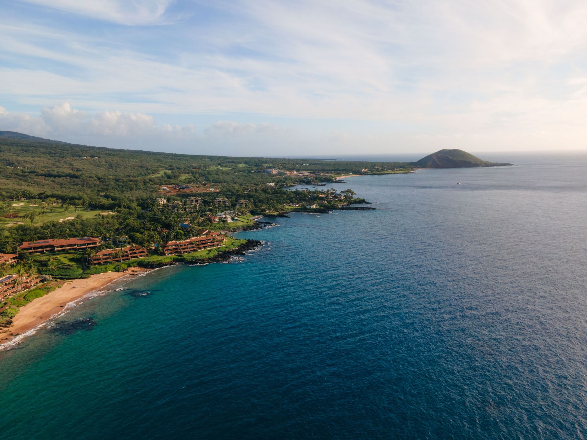 Aerial view of a tropical coastline with blue ocean, sandy beach, and green hills under a cloudy sky