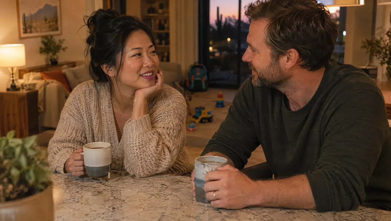 Parents of young kids sitting together at their kitchen island after bedtime, talking over mugs of tea, toys in the soft background