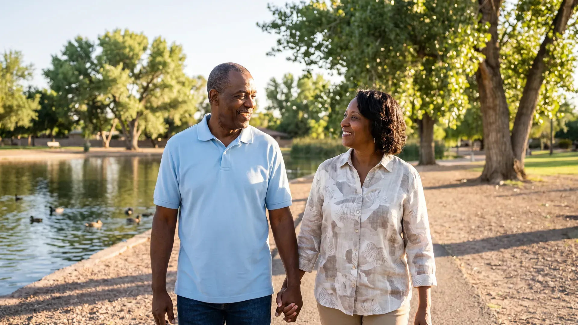 Older married couple strolling arm in arm beside a calm park pond with ducks and tall shade trees, smiling at each other