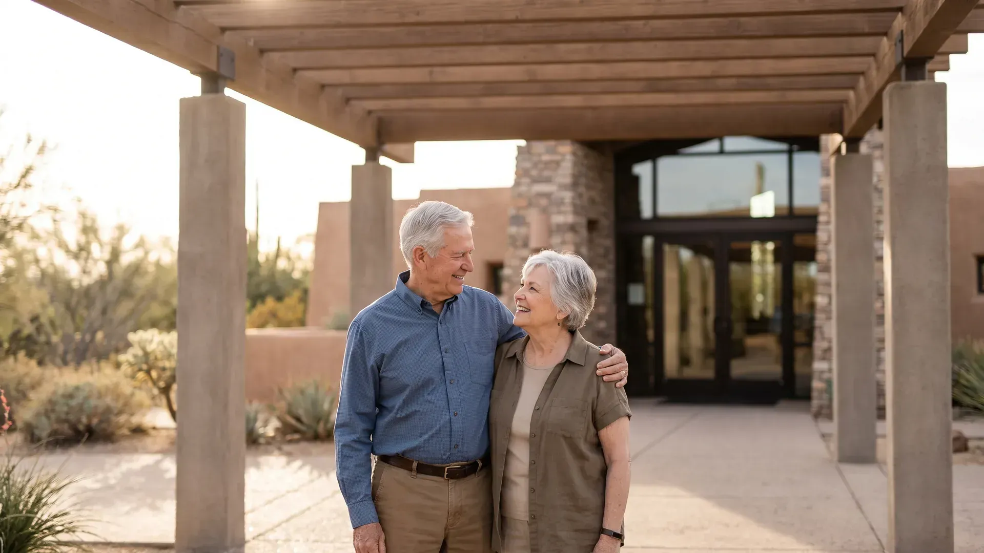 Older married couple standing together under a shaded walkway outside a modern building, his arm around her shoulders, both smiling warmly