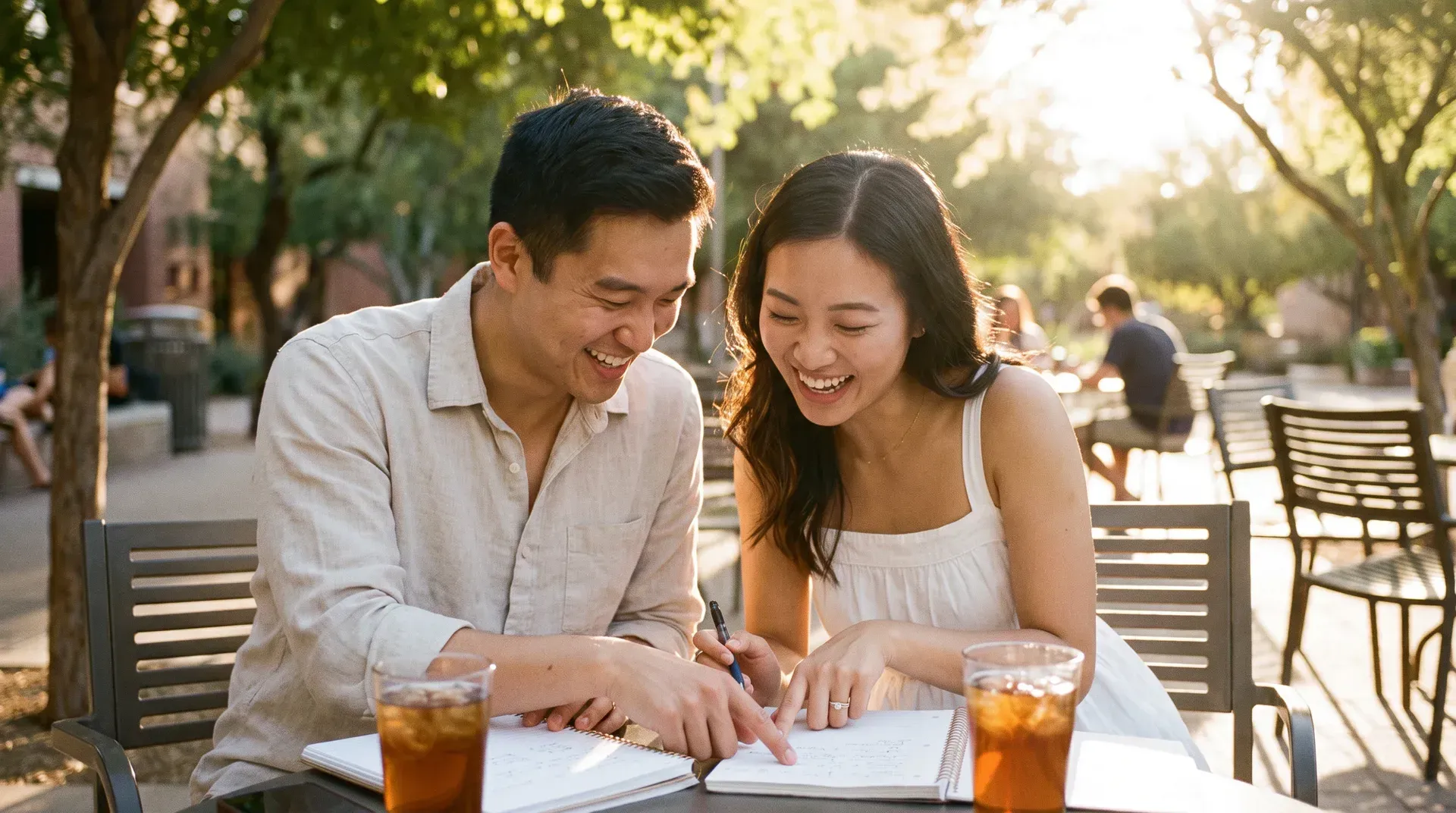 Young married couple in their late twenties sitting at an outdoor campus table with notebooks and iced drinks, planning together and laughing