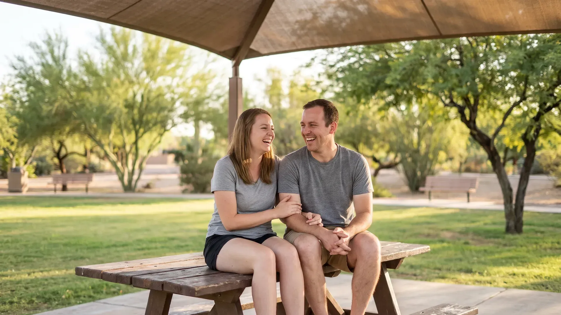 White married couple in their early thirties sitting on a picnic table under a shaded ramada in a green park, shoulders touching, laughing together
