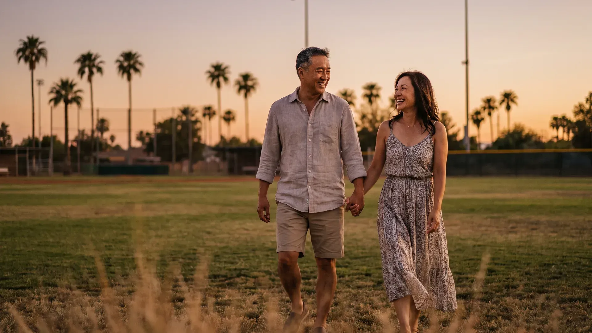 Asian married couple in their mid forties walking hand in hand along the grass edge of a large open green field at sunset, smiling at each other