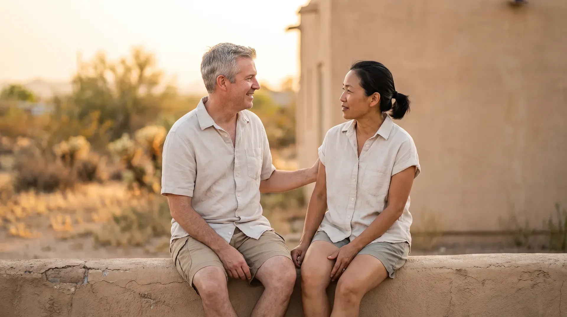 Married couple in their early fifties sitting on a low wall outside a plain office building, turned toward each other, one reaching a hand across