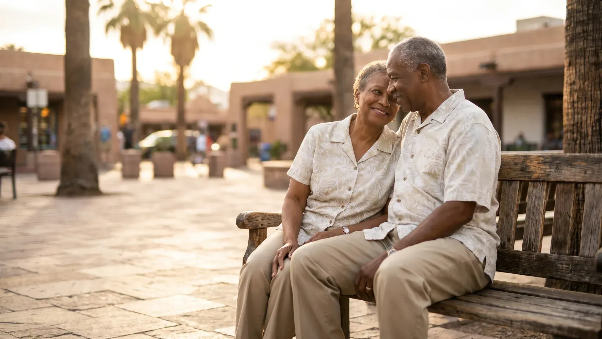Black married couple in their seventies sitting together on a bench in a small plaza lined with palm trees, sharing a warm quiet smile