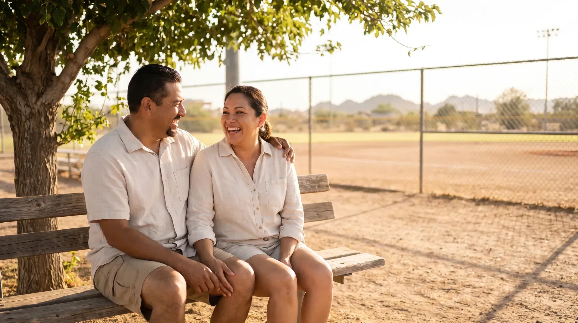 Married couple in their forties sitting side by side on a shaded bench beside a baseball field in the Arizona sun, turned toward each other and laughing