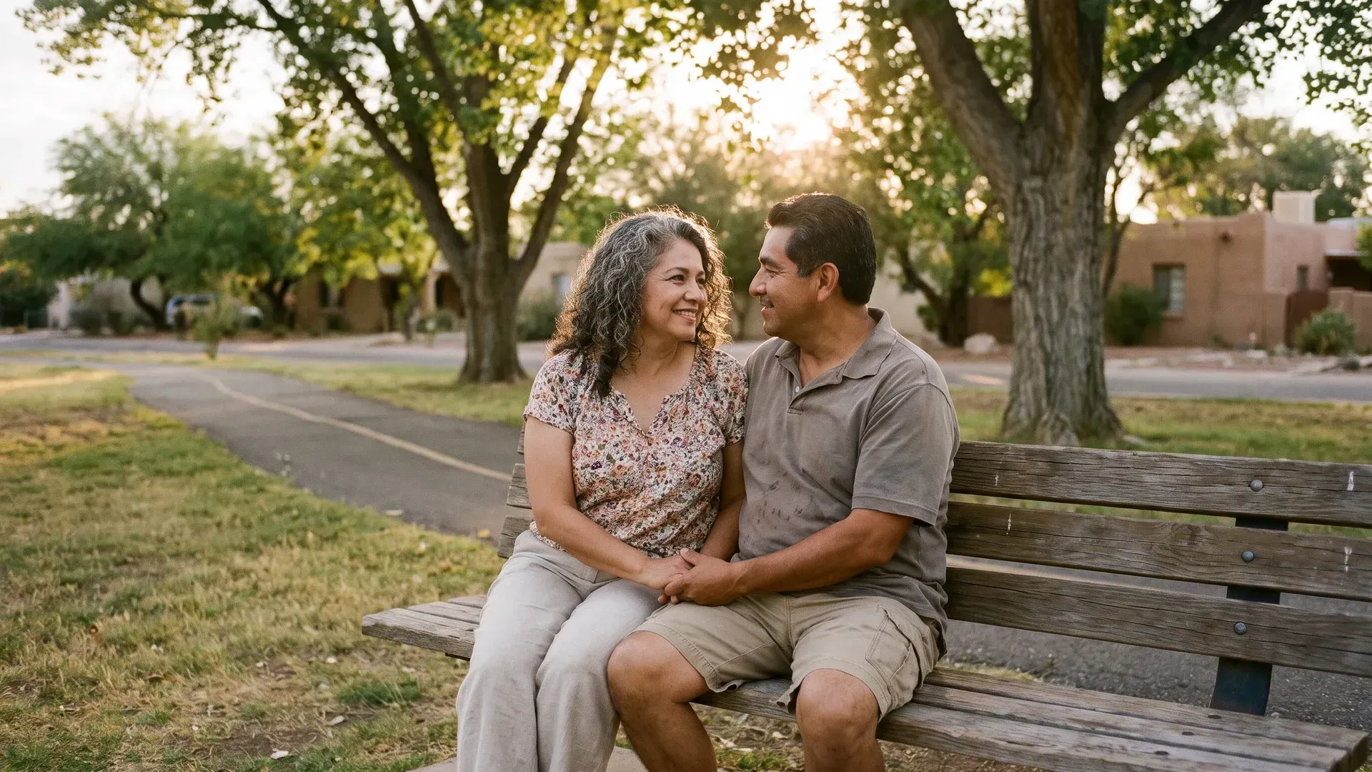 Latino married couple in their early fifties sitting on a park bench under mature shade trees in the evening, turned toward each other in an unhurried honest conversation