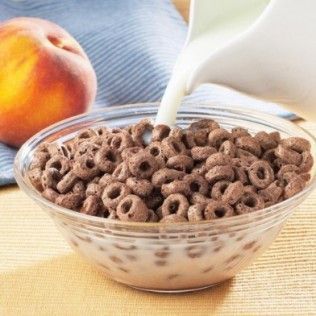 A bowl of chocolate cereal being poured with milk, peach in the background.