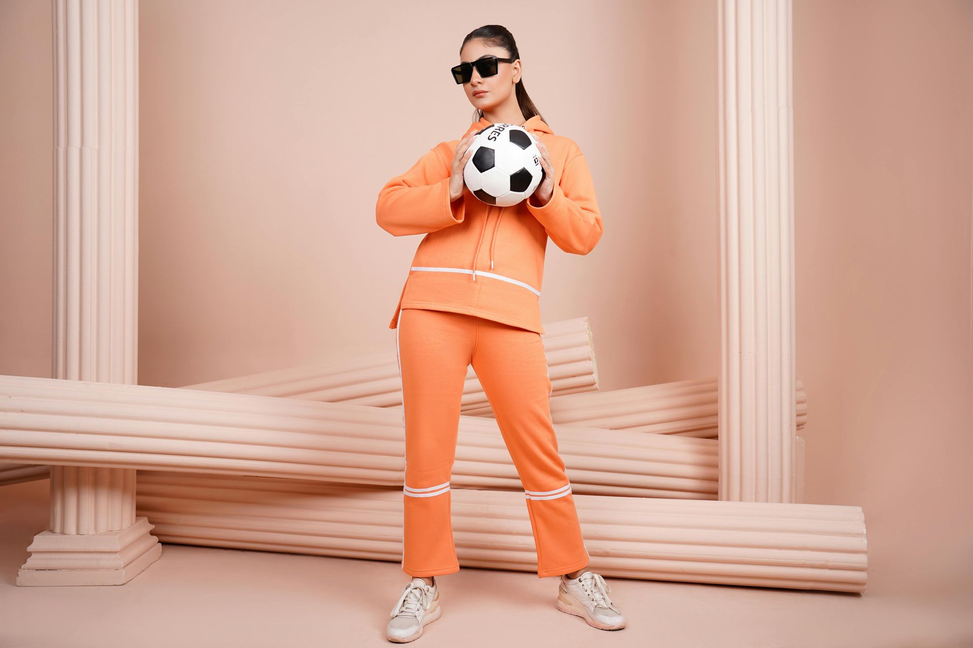 Woman in orange sweatsuit, holding a soccer ball, sunglasses on. Set in front of Roman pillars.