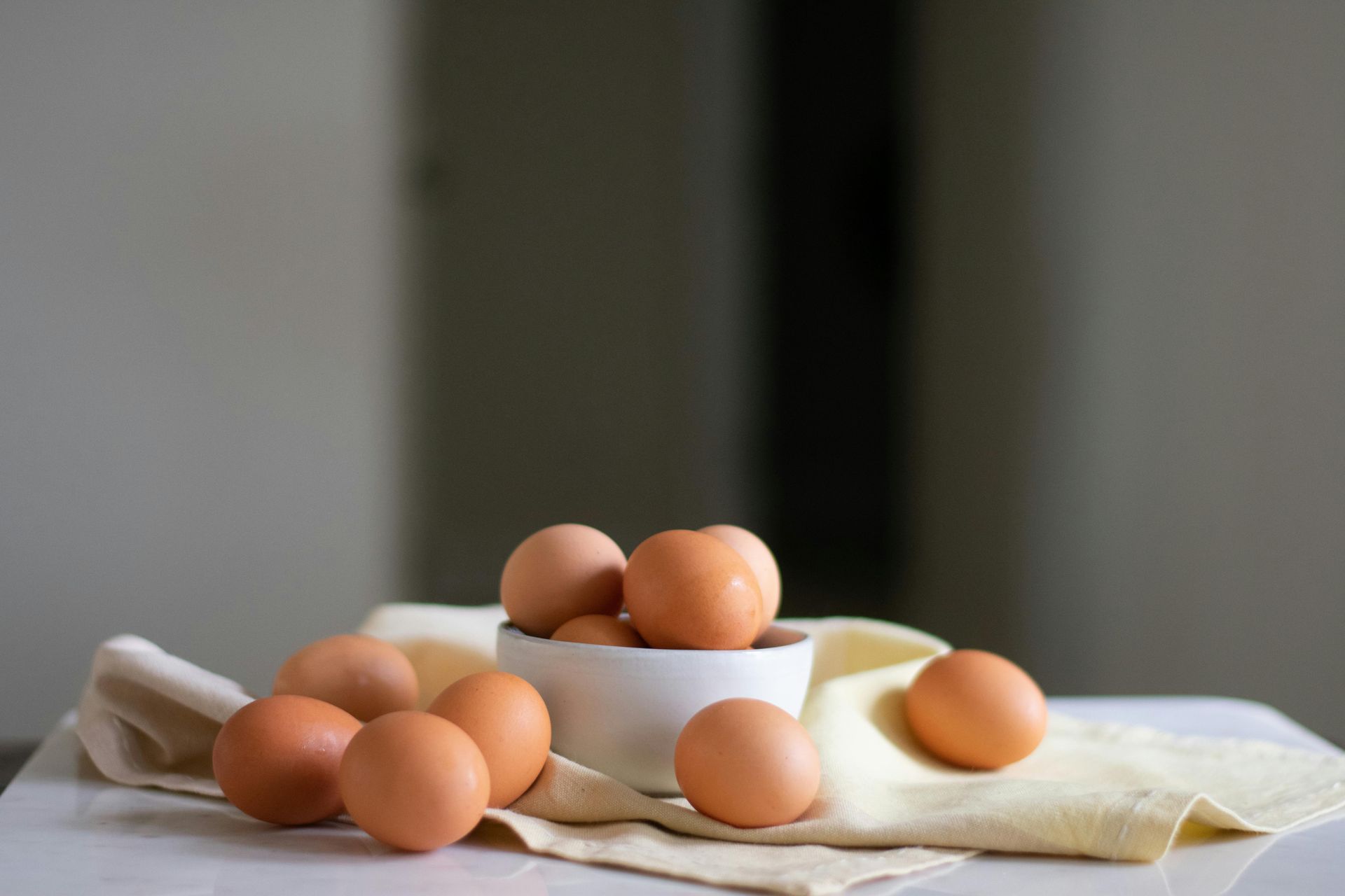 Brown eggs in and around a white bowl, on a light yellow cloth, against a blurred background.