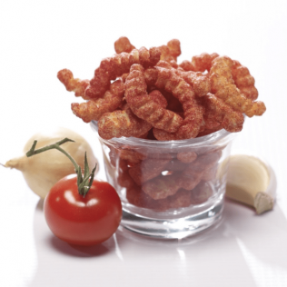Bowl of red, curly-shaped chips next to a tomato, onion, and garlic clove.