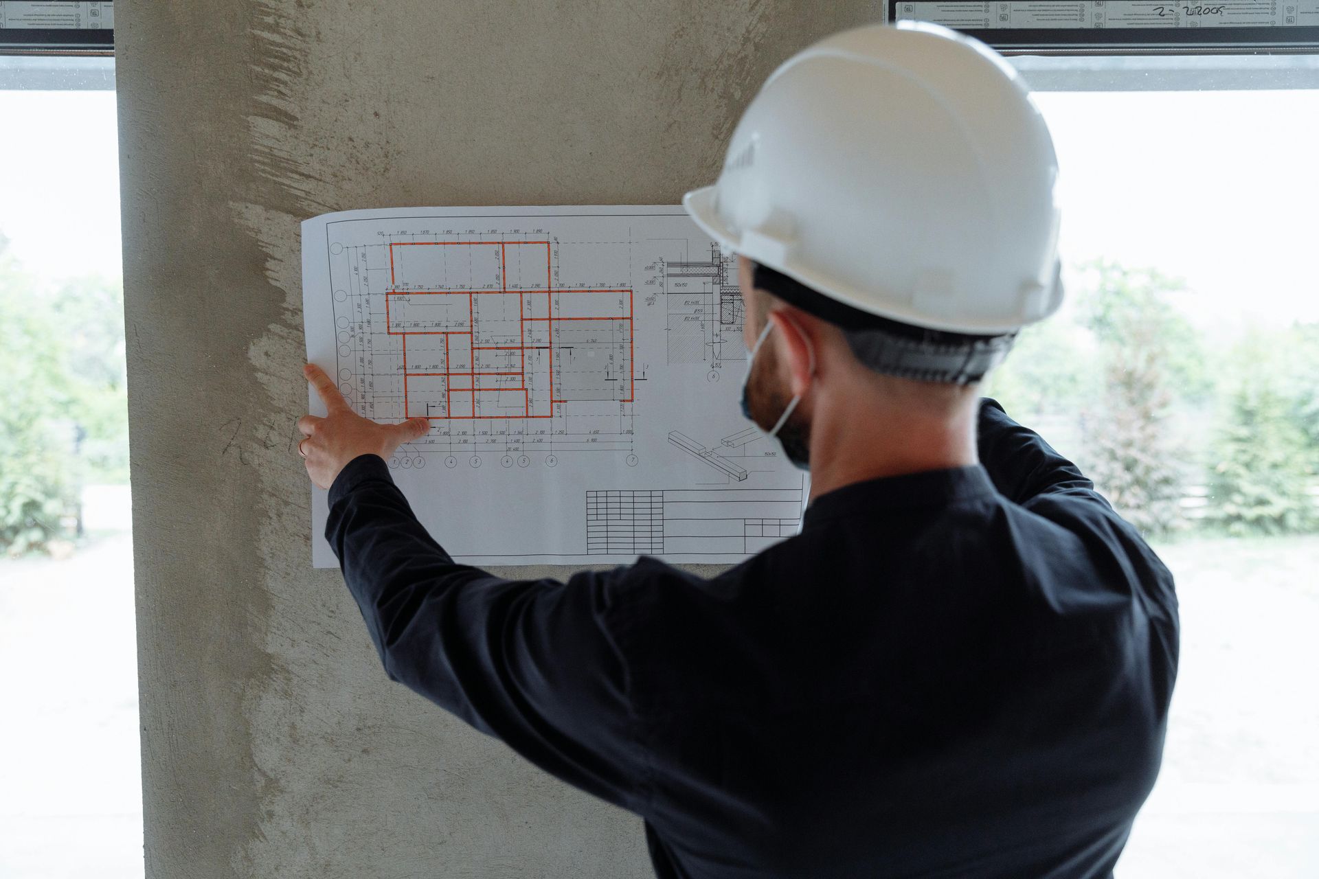 A person in a white hard hat and black shirt holds a structural floor plan against a concrete column at a construction site.