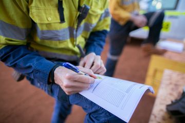 A person in high-visibility work gear writes on a paper form while resting it on their knee outdoors.