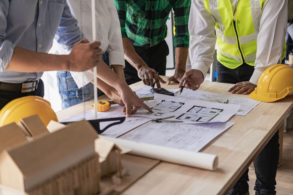 A team of construction professionals reviews architectural floor plans on a wooden table alongside a model house.