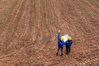 Two people in blue uniforms and yellow hard hats stand on a plowed field, holding a map and pointing at the horizon.