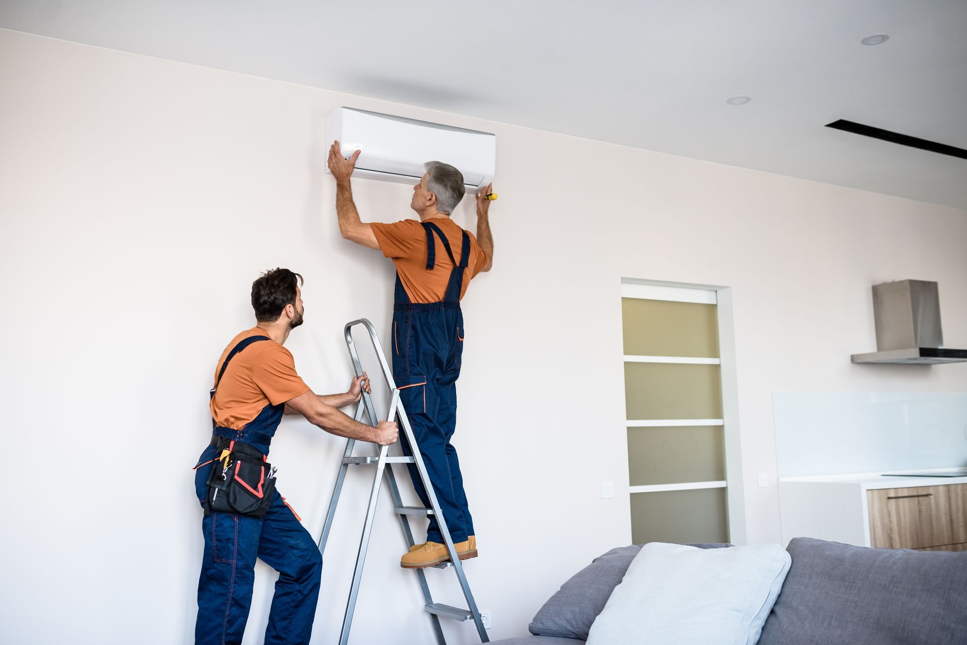 Two workers in orange shirts install an air conditioner above a ladder in a bright room.